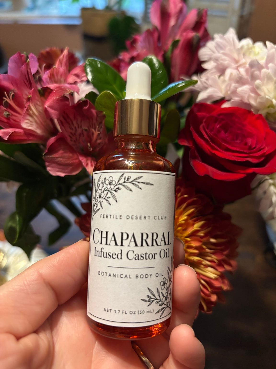 Chaparral-infused castor oil bottle displayed in front of colorful flowers